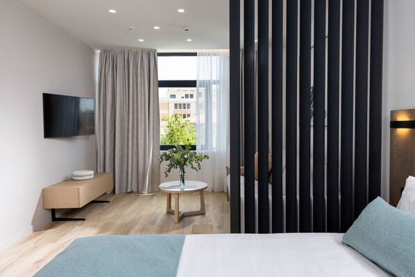 Una habitación de hotel moderna con una cama, una mesita de noche, un televisor montado en la pared, una ventana con cortinas y vistas a edificios.