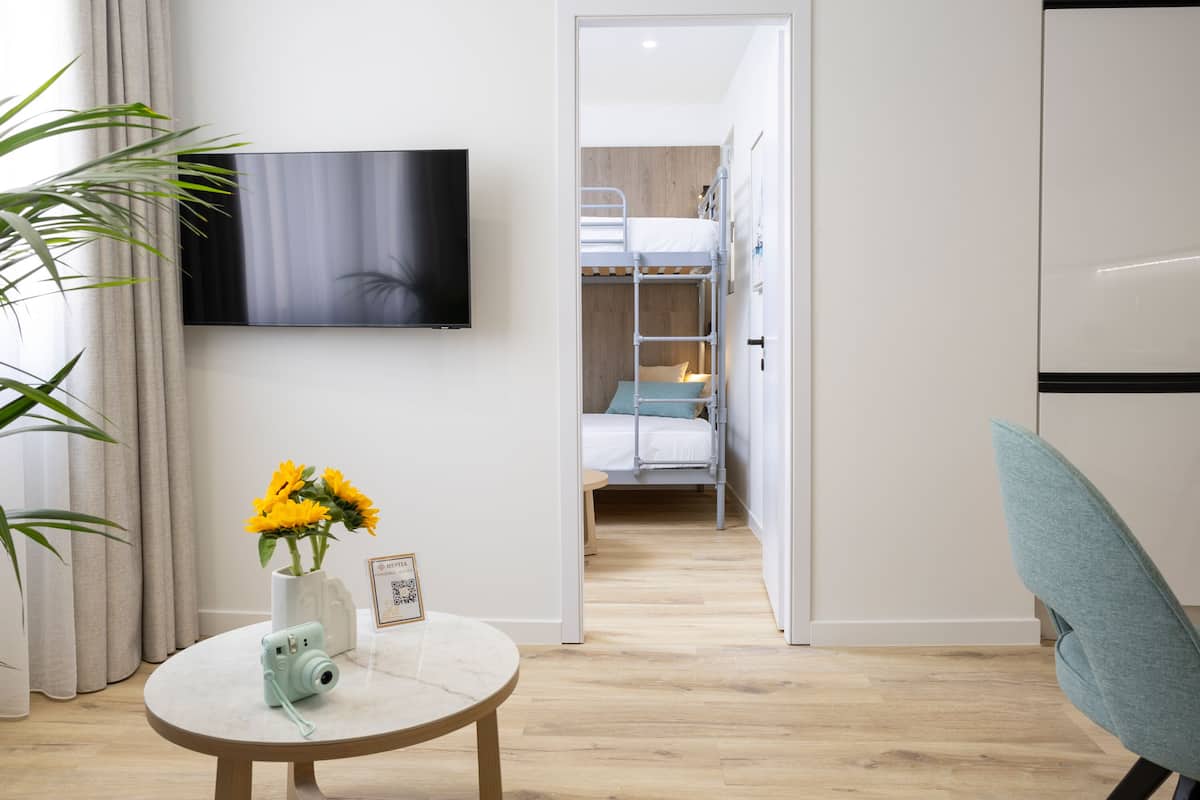 A modern hotel room with a TV mounted on the wall, a bunk bed, a small table with a camera and flowers, and a chair.