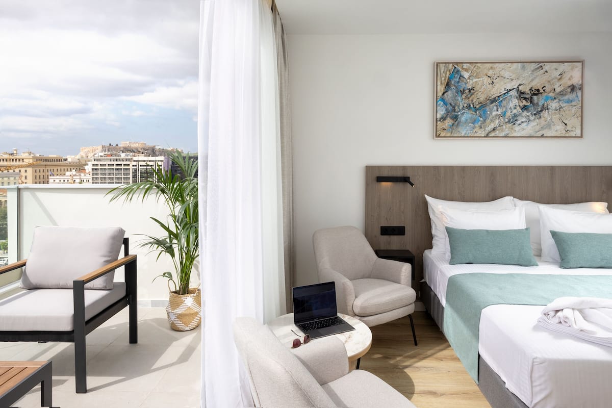 A modern hotel room with a balcony, a bed with white and teal bedding, a laptop on a small table, and a city view.