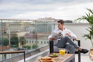 A man reading a newspaper on a balcony with a city view, a table with food and drinks, and a potted plant. - Hestia - Asklipiou 9 (Athens)
