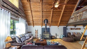 A cozy living room with a wooden ceiling, a fireplace, leather sofas, and a large window with curtains. - Cabin at the Forest Edge | Hot tub, pet friendly (Packwood)
