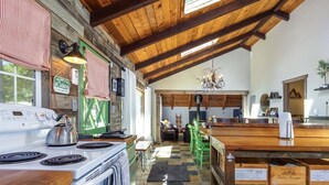 A rustic kitchen with a wooden ceiling, a stove, and a dining area with green chairs. - Cabin at the Forest Edge | Hot tub, pet friendly (Packwood)