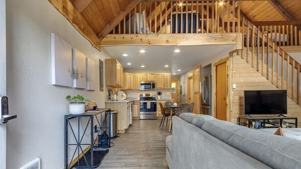 A modern kitchen with wooden cabinets, a dining area with a grey sofa, and a staircase leading to a loft. - Hygee Haus | Hot Tub, Wifi, Wood stove (Packwood)