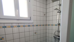 Une salle de bain avec des murs recouverts de carreaux blancs, un sol carrelé de bleu, une douche équipée d’un pommeau de douche à main et une fenêtre au-dessus du lavabo.
