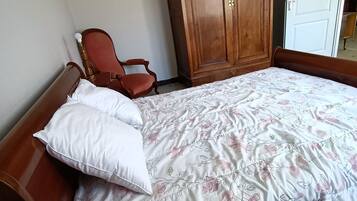 Ein Schlafzimmer mit großem Holzschrank, einem Einzelbett mit Blumenmuster auf der Bettwäsche, einem Holzstuhl und einer weißen Tür im Hintergrund.