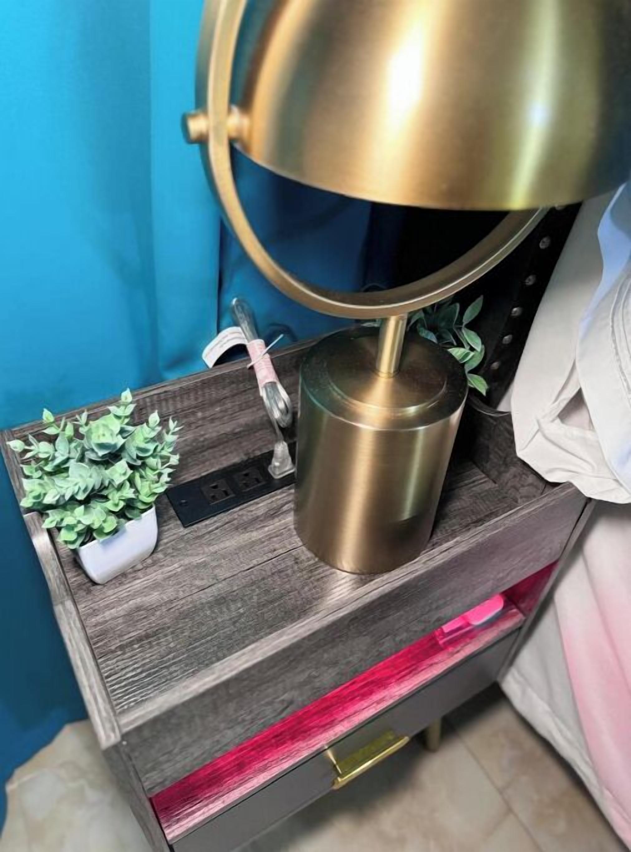 A bedside table with a brass lamp, a small potted plant, and a power outlet.