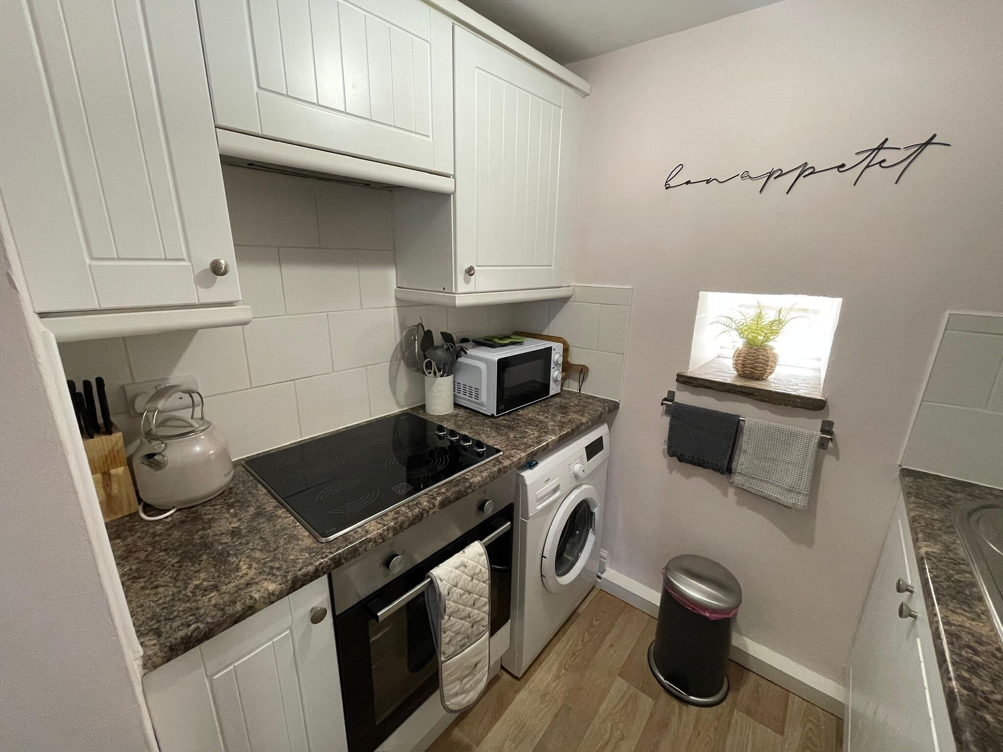 A compact kitchen with white cabinets, a microwave, a washing machine, and a small window with a plant.