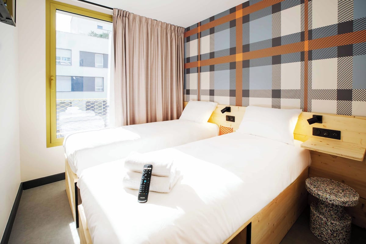 A hotel room with a bed, a window with curtains, a plaid-patterned wall, and a small stool.