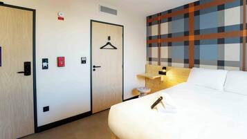 A hotel room with a bed, a bedside table with a lamp, a wall-mounted TV, a door with a hanger, and a wall with a plaid pattern.