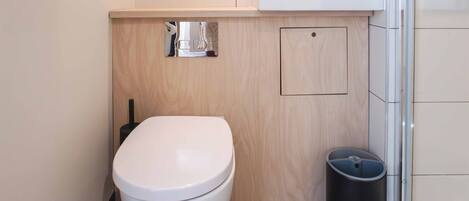 Ein modernes Badezimmer mit Toilette in Weiß, einem schwarzen Mülleimer, einem Holzkosmetikschrank und einer Duschkabine aus Glas.