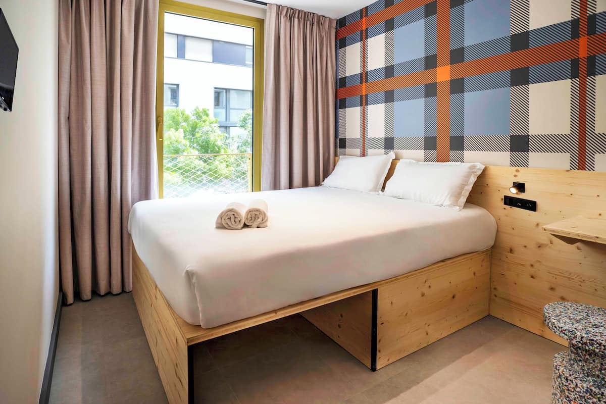 A modern hotel room with a wooden bed, a view of greenery, and a plaid-patterned wall.