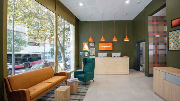 A reception area with a wooden desk, a green chair, and a brown sofa. There are large windows with a view of the street and a bus. - easyHotel Marseille Euromed (Marseille)