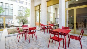 An outdoor patio area with red metal tables and chairs, a brick floor, and large windows overlooking a parking area. - easyHotel Marseille Euromed (Marseille)