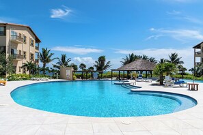 A large, clear swimming pool with lounge chairs and a gazebo, surrounded by palm trees and a building with balconies. - Oceanfront Bliss: Luxurious 2BR Condo in Ambergris Caye! (San Pedro Town)