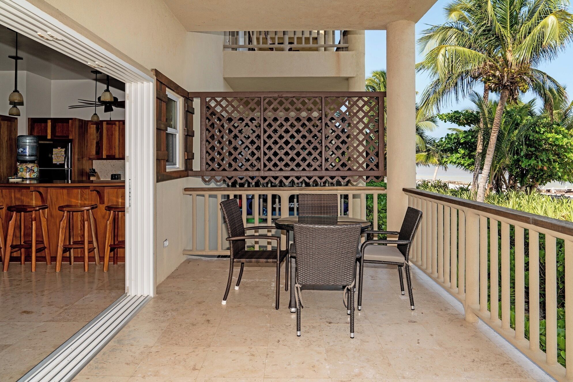A balcony with a dining table and chairs, a kitchen area with bar stools, and a view of palm trees.