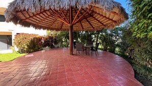 A covered outdoor seating area with a thatched roof, surrounded by lush greenery and a paved walkway.