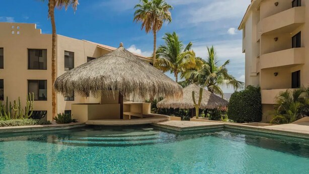 A swimming pool with clear blue water, surrounded by palm trees and thatched umbrellas.