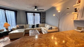 A modern living room with a beige sectional sofa, a coffee table, and a kitchen area with granite countertops and a white refrigerator.