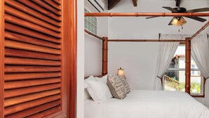 Un lit à baldaquin en bois, agrémenté de draps blancs et de coussins décoratifs, un ventilateur de plafond et une vue sur la verdure environnante.