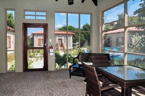 Una habitación con una mesa de vidrio y sillas de mimbre, ventanas amplias con vistas a una piscina y edificios.