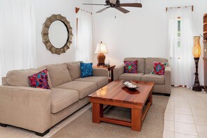 A living room with two beige sofas, colorful pillows, a wooden coffee table, a floor lamp, and a ceiling fan.