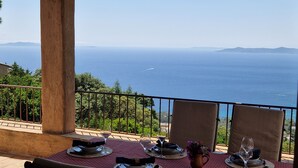 A dining table set with plates, glasses, and a vase of flowers, overlooking a coastal view with the sea and distant mountains.