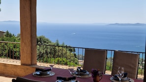 Mesa de jantar posta com pratos, copos e um vaso de flores, com vista para a costa, o mar e montanhas ao longe.