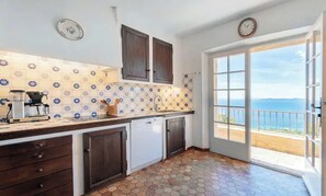 Uma cozinha com painel de azulejos, armários de madeira, máquina de lavar louça e vista para o mar através de uma porta de vidro.