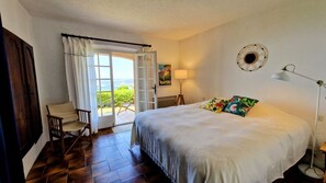 Quarto com cama, cadeira, escrivaninha, luminária e vista por uma porta aberta.