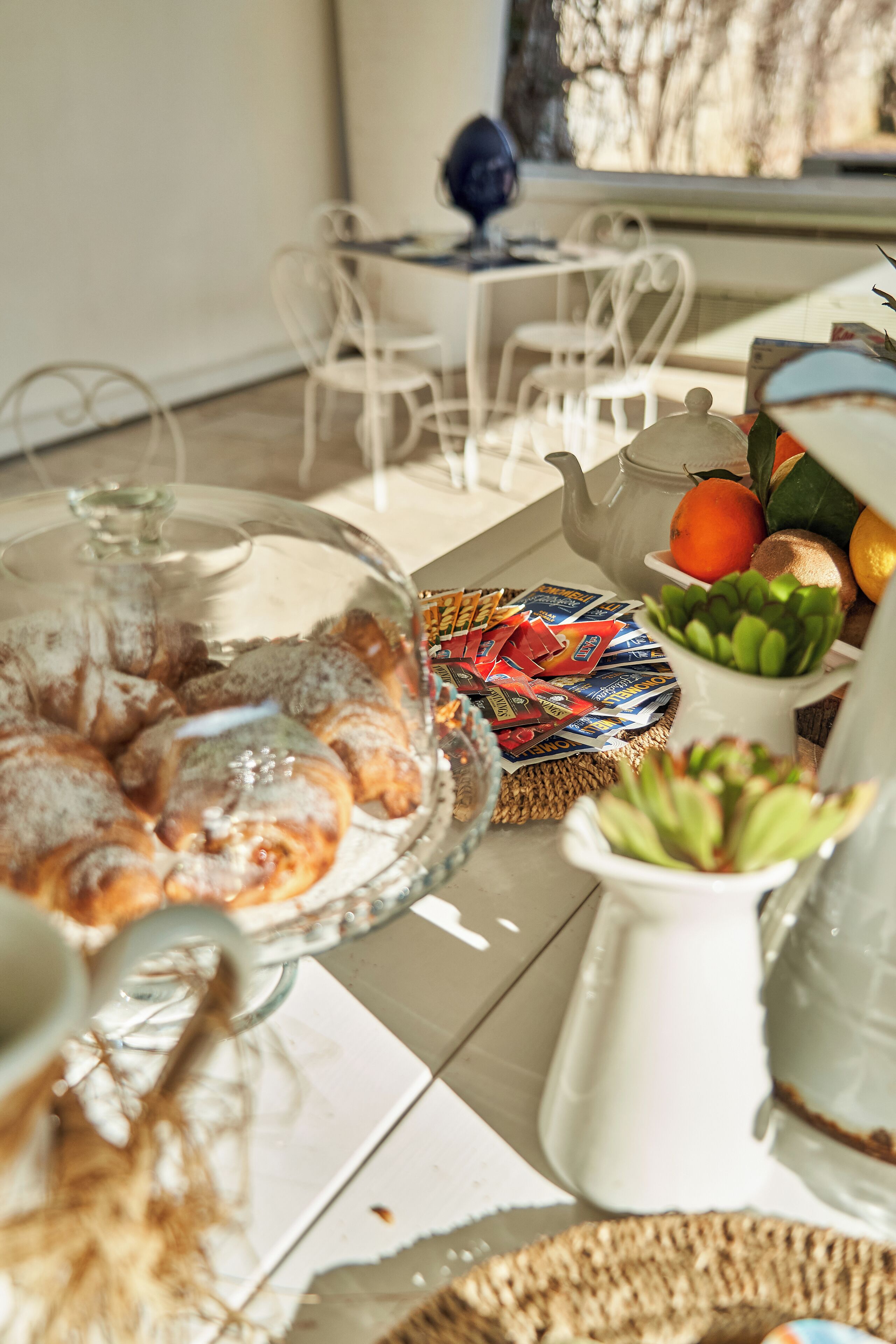 A dining table with a glass-covered pastry display, a teapot, and various food items.