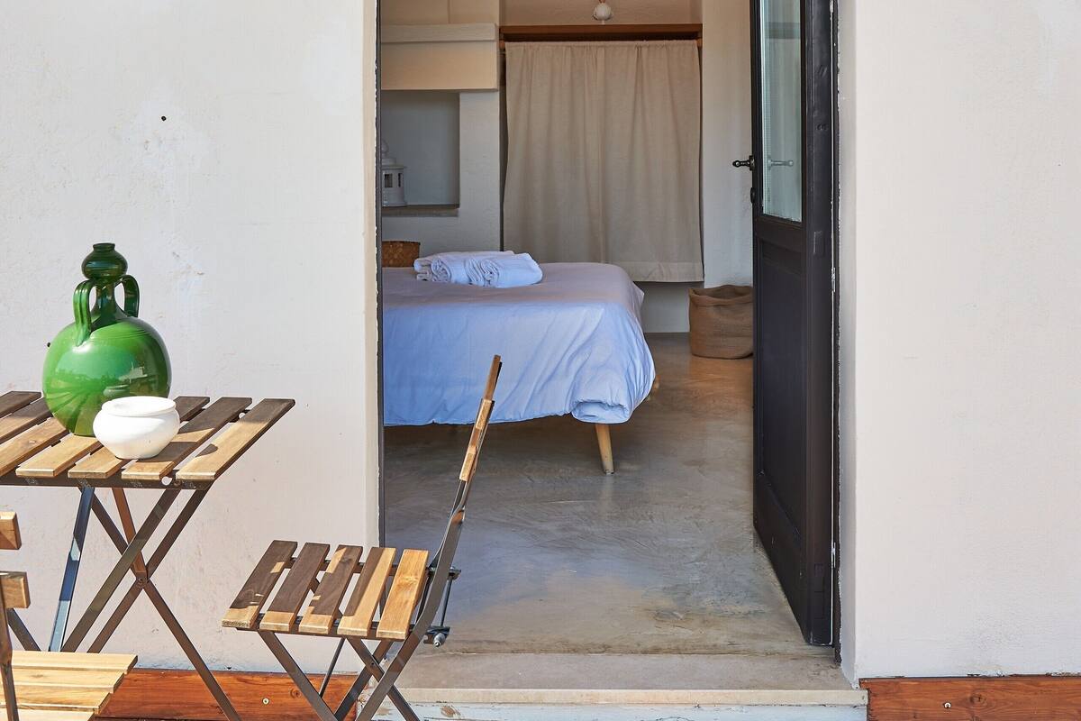 Una terraza de madera con una mesa y sillas, un contenedor verde, y un dormitorio con cama, cortinas y una lámpara colgante.