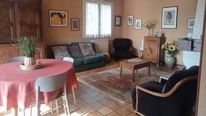 A living room with a sofa, chairs, a coffee table, and a dining table. There are framed pictures on the walls and a window with curtains.