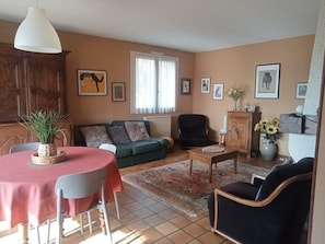 Un salon comprenant un canapé, des fauteuils, une table basse et une table à manger. Les murs sont ornés de tableaux encadrés et il y a une fenêtre avec des rideaux.