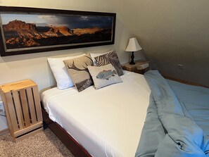 Cama bem arrumada com roupa de cama branca e manta azul, com uma mesinha de cabeceira de madeira com abajur e um pequeno banquinho de madeira.