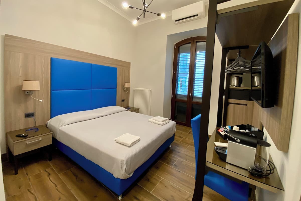 A modern hotel room with a large bed, a bedside table, a nightstand, a mirror, a wardrobe, and a kitchenette area.