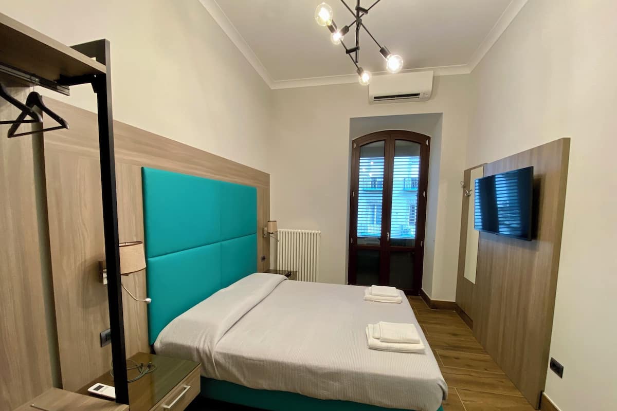 A modern hotel room with a teal accent wall, a bed with white linens, a TV mounted on the wall, and a wooden floor.
