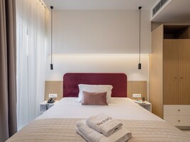 Una cama bien tendida con cabecera roja, una mesita de noche con un teléfono y un armario de madera.