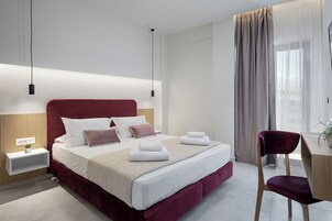 A modern hotel room with a large bed, a bedside table with a lamp, a desk with a chair, and a window with curtains.