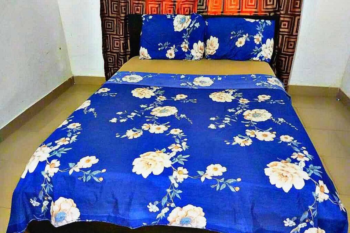A bed with a blue floral bedspread and matching pillow, a patterned headboard, and a plain wall.