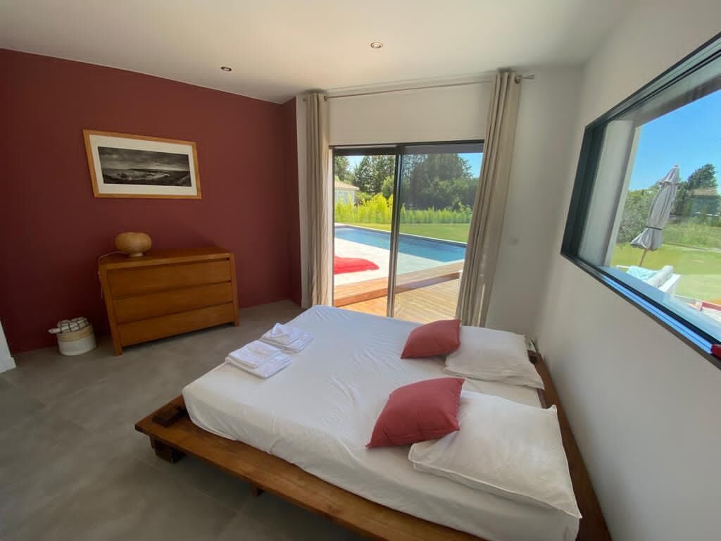 A bedroom with a bed, a wooden dresser, a framed picture on the wall, and a view of a swimming pool through a sliding glass door.