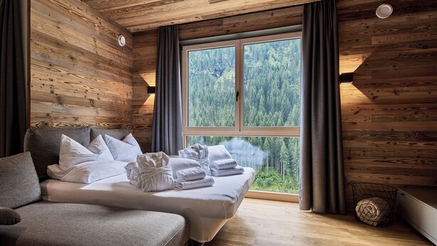 A wooden cabin room with a bed, a sofa, and a large window overlooking a forest. - sGmiatli – Hideaway Apartments (Kappl)