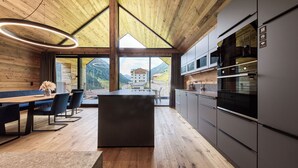 A modern kitchen with wooden ceiling and large windows, a dining area with a wooden table and chairs, and a view of mountains. - sGmiatli – Hideaway Apartments (Kappl)