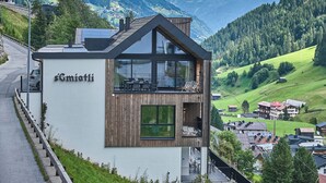 A modern building with a wooden facade and large windows, named "Sgiatli," overlooking a green valley with houses and mountains in the background. - sGmiatli – Hideaway Apartments (Kappl)