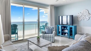 A spacious living room with a large sliding glass door leading to a balcony with a view of the ocean. There is a flat-screen TV mounted on a blue cabinet, a ceiling fan, and a decorative wall piece. - Beachfront End-Unit, Sunset Views! Beach Service! 4th Floor Shores of Panama (Panama City Beach)