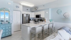 A modern kitchen with a central island, white cabinetry, stainless steel appliances, and a view to the outside. - Beachfront End-Unit, Sunset Views! Beach Service! 4th Floor Shores of Panama (Panama City Beach)
