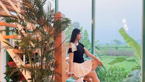 A woman sitting by a large window with a view of a green landscape. - Room in Chalet - Trang An Ao Dai Homestay - Wooden House With Field View and Private Bathroom (Hoa Lu)