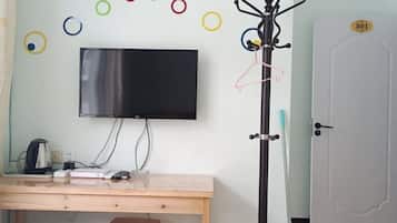 Una habitación con una televisión de pantalla plana montada en la pared, un escritorio de madera con una tetera y un vaso, un perchero con ropa y una puerta con placa de identificación.