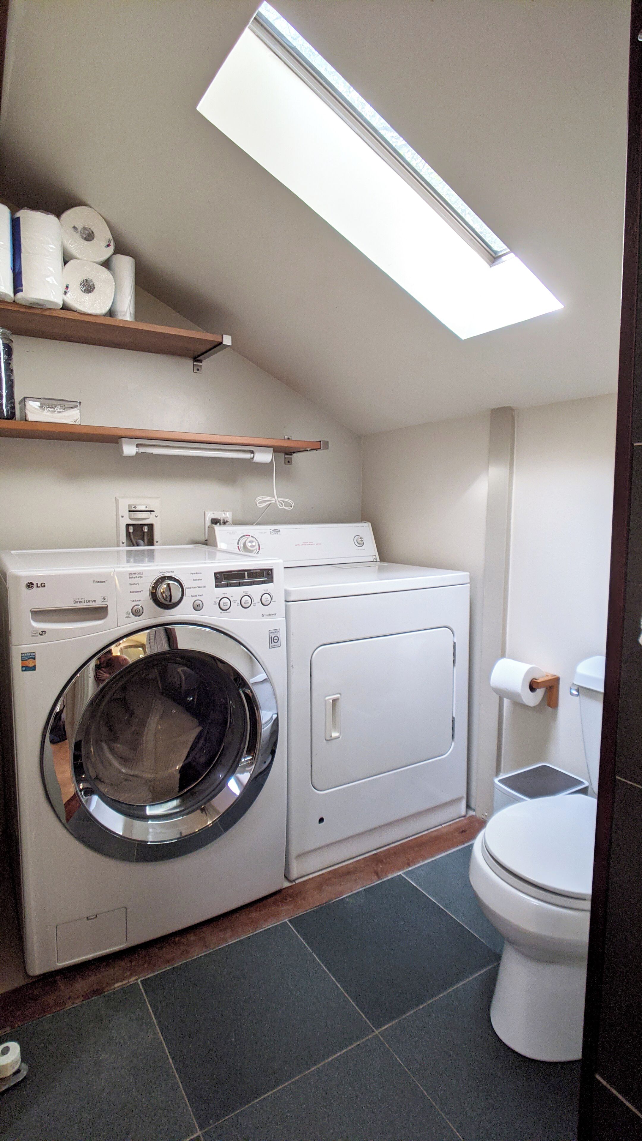A compact laundry room with a front-loading washer and dryer, a toilet, and a skylight above.