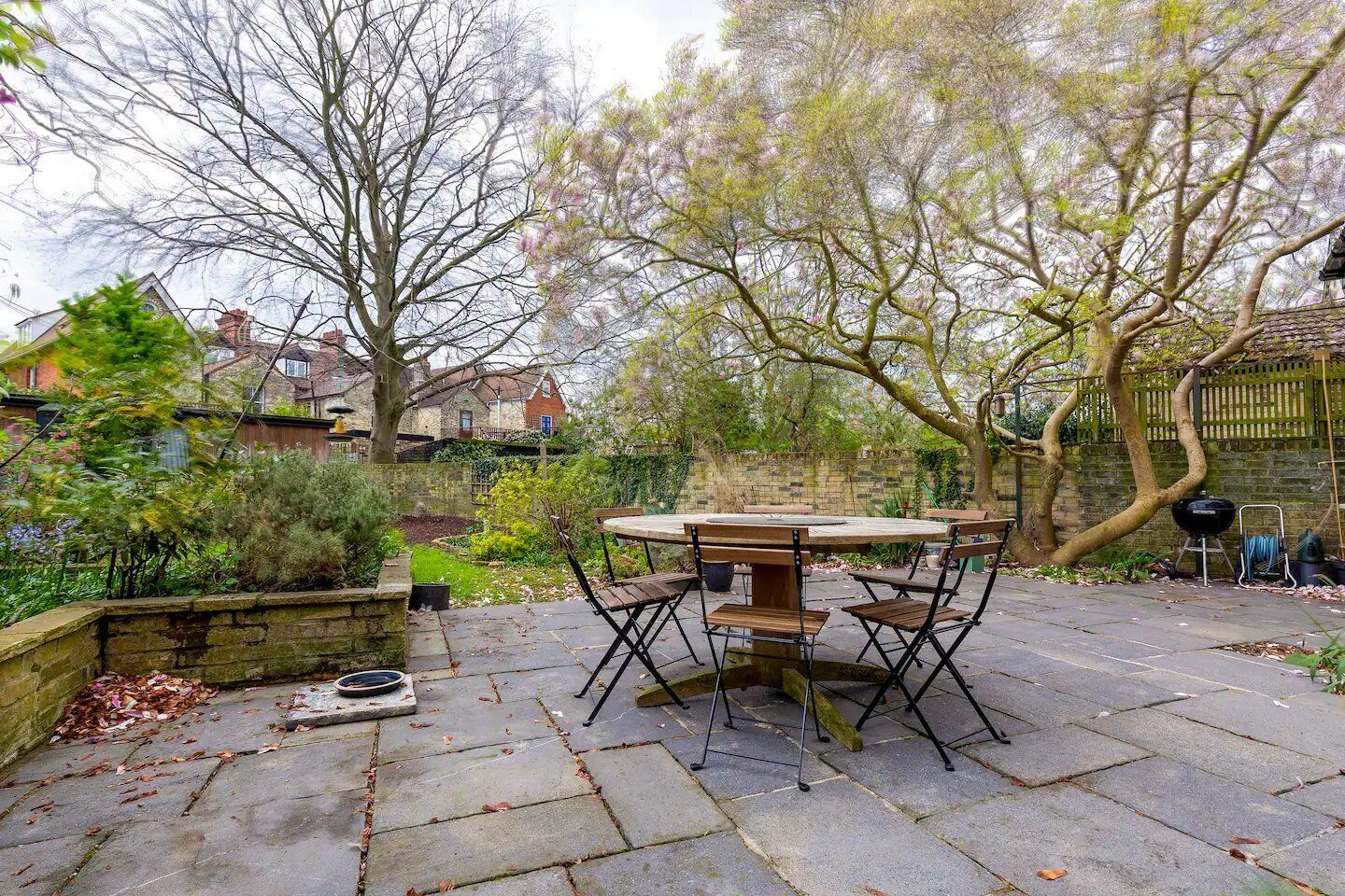 An outdoor patio with a round table and chairs, surrounded by stone paving, trees, and a brick wall.