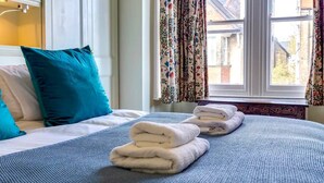 Habitación de hotel con cama, ropa de cama azul, almohadas color turquesa y una ventana con cortinas florales.